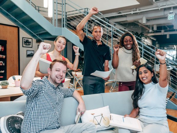 Five happy multi-ethnic engineering students in casual dress gathered around a couch with their designs for a bicycle. Their fists are raised in victory for their successful teamwork.